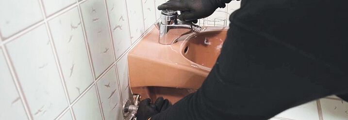Ein Mann mit Handschuhen dreht Wasserzulauf am Waschbecken zu.